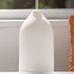 A white essential oil diffuser with misting at the top, sitting on a white surface in front of a window. dōTERRA Laluz Diffuser with Lemon and Lavender