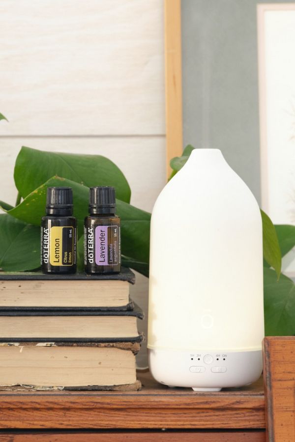 A white LED diffuser lamp sits on a wooden shelf next to two small doTERRA essential oil bottles on a stack of books, with green leaves in the background. dōTERRA Laluz Diffuser with Lemon and Lavender