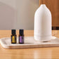 A white essential oil diffuser sits on a light stone tray next to two small amber bottles on a wooden table. dōTERRA Laluz Diffuser with Lemon and Lavender