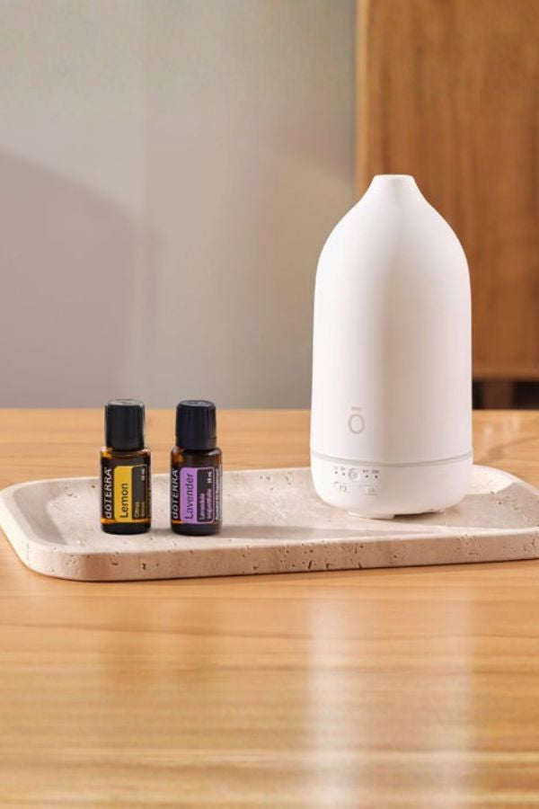 A white essential oil diffuser sits on a light stone tray next to two small amber bottles on a wooden table. dōTERRA Laluz Diffuser with Lemon and Lavender
