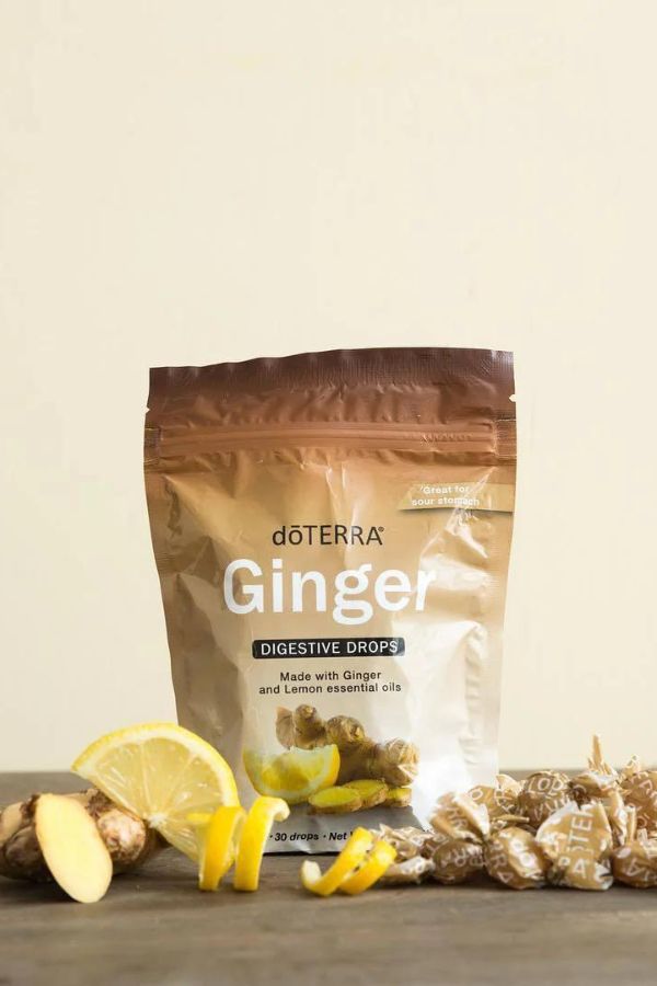 A stand-up pouch of doTERRA Ginger Digestive Drops displayed on a wooden surface with ginger root slices, lemon wedges, and wrapped capsules around it, against a plain beige background. doTERRA Ginger Digestive Drops (2 Pack)