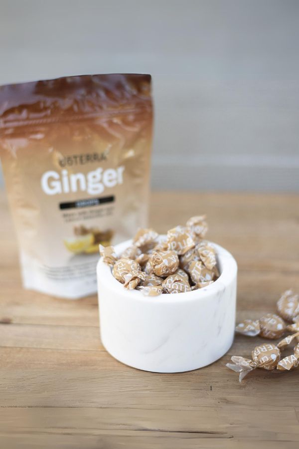 A stand-up pouch of DoTERRA Ginger candy sits on a wooden tabletop behind a small white marble bowl filled with ginger candies, with a few candies scattered nearby.