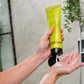 A person's hands squeezing a bright yellow-green shampoo tube under running water in a shower, with white shampoo lather dripping into the palm. doTERRA Salon Essentials Smoothing Conditioner is 