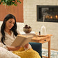 A woman sits in a cozy living room by a fireplace, reading a book wrapped in a chunky mustard yellow knit blanket. A wooden side table beside her holds a white diffuser, a small bowl, and books, with a white brick wall and lit fireplace in the background. dōTERRA Harvest Spice Blend | Canada