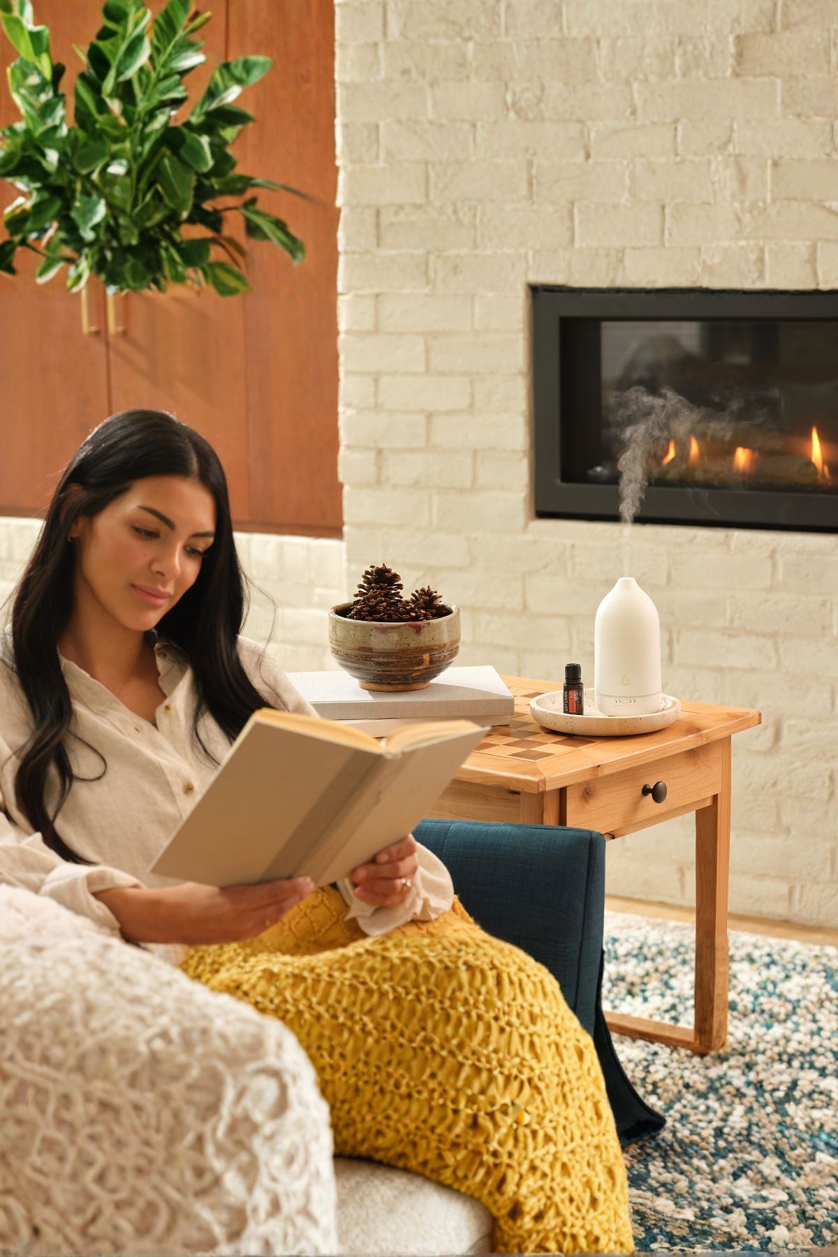A woman sits in a cozy living room by a fireplace, reading a book wrapped in a chunky mustard yellow knit blanket. A wooden side table beside her holds a white diffuser, a small bowl, and books, with a white brick wall and lit fireplace in the background. dōTERRA Harvest Spice Blend | Canada