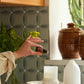 A hand holds a small amber essential oil bottle in a kitchen setting with patterned gray-green tile backsplash, ceramic pots, a white diffuser, and a candle on a woven placemat. dōTERRA Harvest Spice Blend | Canada