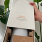 A hand holding the lid of a white Laluz diffuser box, revealing a white diffuser bottle and a brown instruction card inside a brown foam insert, with green plant leaves in the background. dōTERRA Laluz Diffuser with Lemon and Lavender