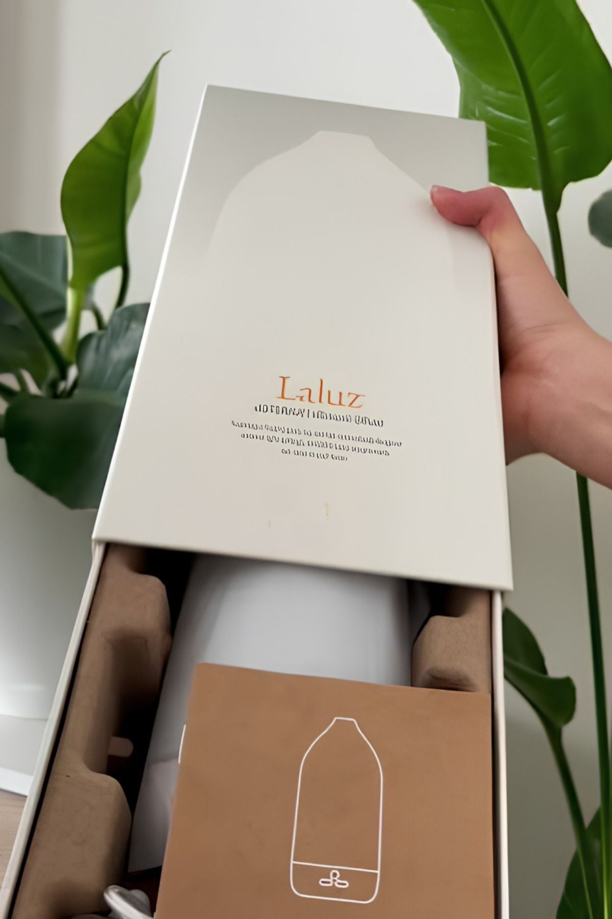 A hand holding the lid of a white Laluz diffuser box, revealing a white diffuser bottle and a brown instruction card inside a brown foam insert, with green plant leaves in the background. dōTERRA Laluz Diffuser with Lemon and Lavender