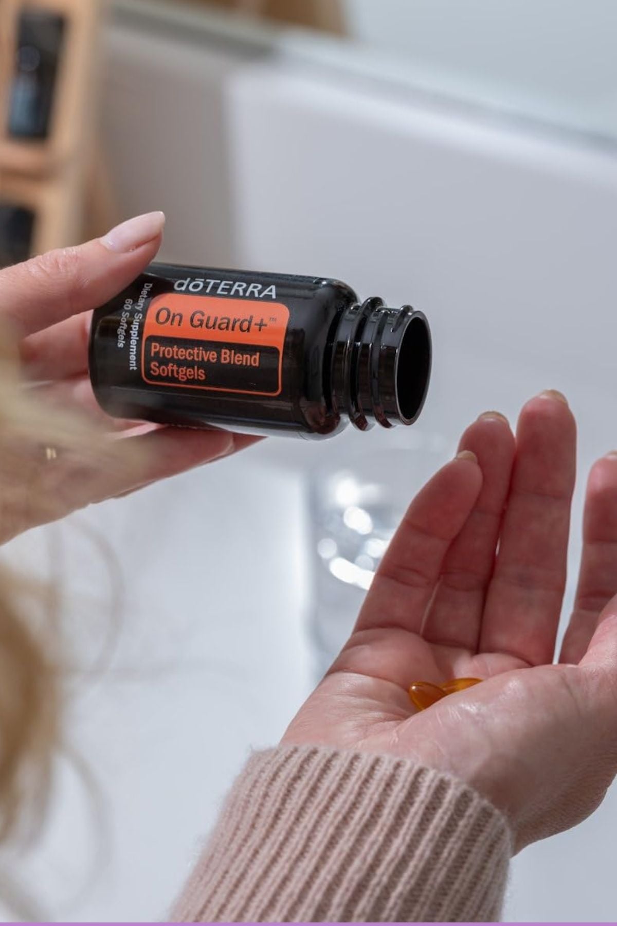 A person holds an open bottle of doTERRA On Guard+ Protective Blend Softgels over their palm, with a small amber softgel visible in their other hand, set against a softly blurred bathroom background. doTERRA On Guard Softgels (2 Pack)