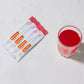 Four white and orange MucinPower liquid supplement packets are arranged on a light marble surface next to a clear glass containing a bright red liquid. dōTERRA MetaPWR Recharge Electrolytes (Wild Strawberry)
