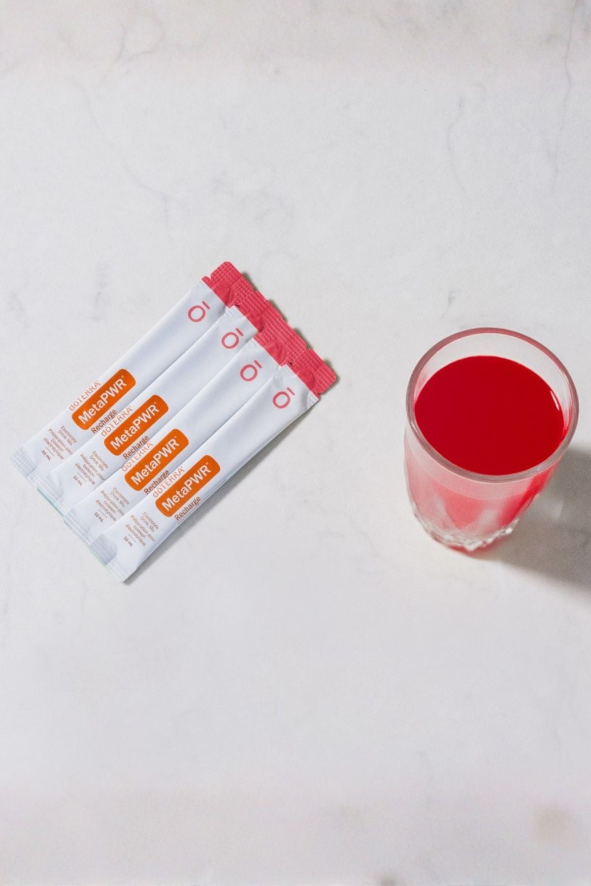 Four white and orange MucinPower liquid supplement packets are arranged on a light marble surface next to a clear glass containing a bright red liquid. dōTERRA MetaPWR Recharge Electrolytes (Wild Strawberry)