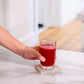 A hand holding a clear glass filled with bright red juice, resting on a square concrete coaster on a white marble countertop in a bright kitchen setting. dōTERRA MetaPWR Recharge Electrolytes (Wild Strawberry)