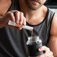 A man in a gray tank top mixes a red liquid into a clear shaker bottle with a black sleeve, holding a white and orange MicroPlenty packet above the bottle. dōTERRA MetaPWR Recharge Electrolytes (Wild Strawberry)