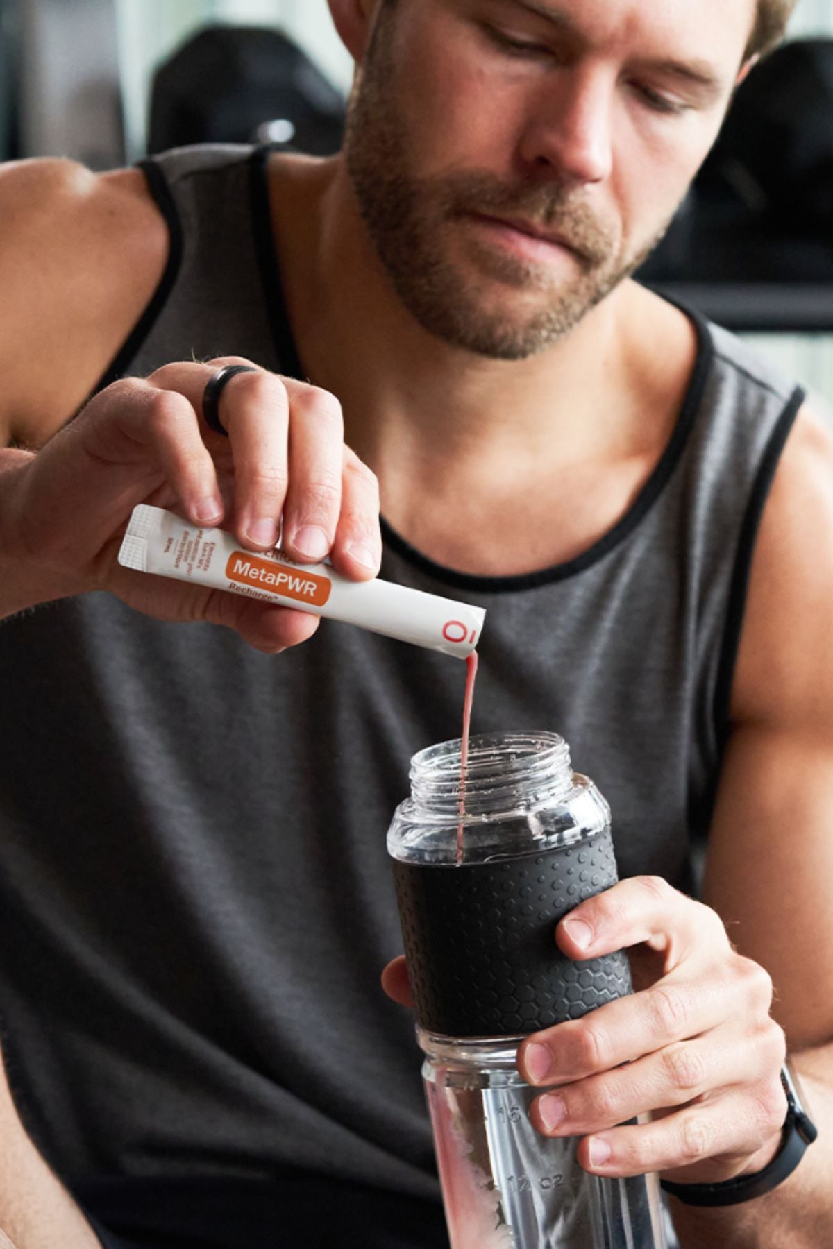 A man in a gray tank top mixes a red liquid into a clear shaker bottle with a black sleeve, holding a white and orange MicroPlenty packet above the bottle. dōTERRA MetaPWR Recharge Electrolytes (Wild Strawberry)