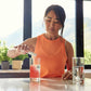 A woman in an orange sleeveless top stands at a kitchen counter pouring a pink drink into a glass, with two clear glasses and a bright window view of mountains in the background. dōTERRA MetaPWR Recharge Electrolytes (Wild Strawberry)