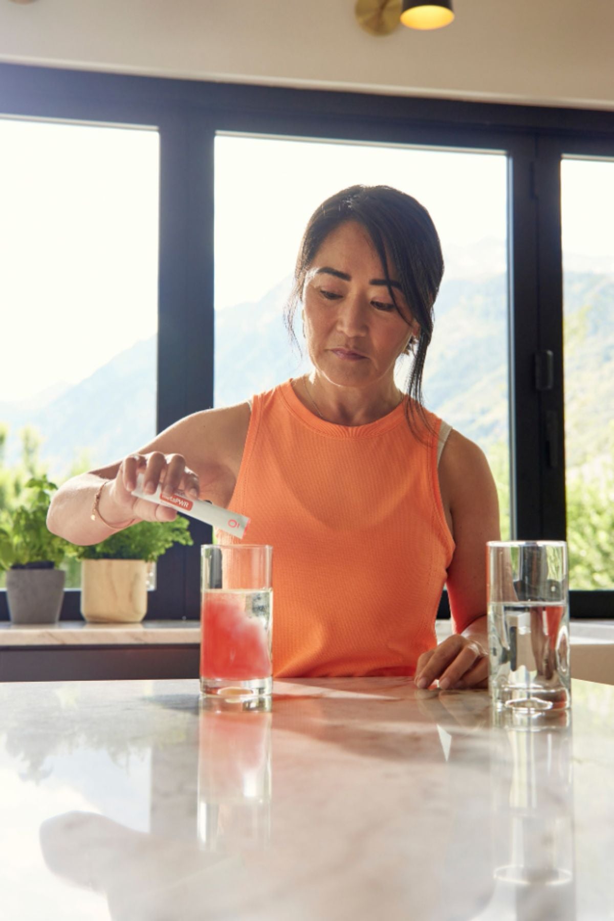 A woman in an orange sleeveless top stands at a kitchen counter pouring a pink drink into a glass, with two clear glasses and a bright window view of mountains in the background. dōTERRA MetaPWR Recharge Electrolytes (Wild Strawberry)